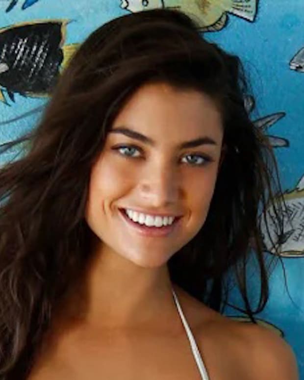 Lauren Mellor stands in front of a blue wall with painted fish and smiles at the camera in a white string bikini.